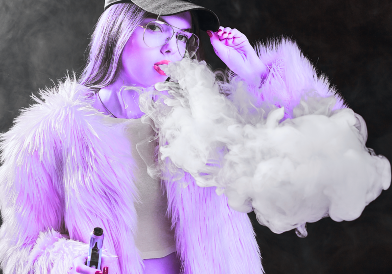 young woman in purple jacket blowing smoke from live resin disposable vape