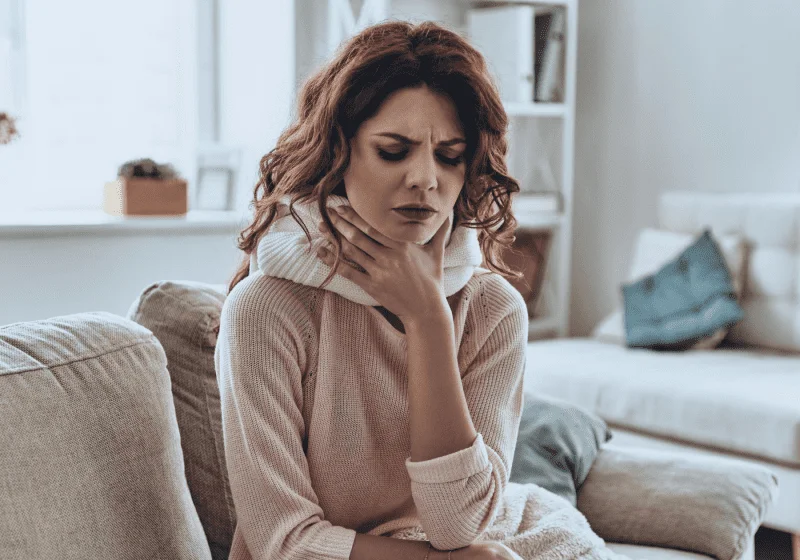 woman with red hair clutching her sore throat while sitting on couch