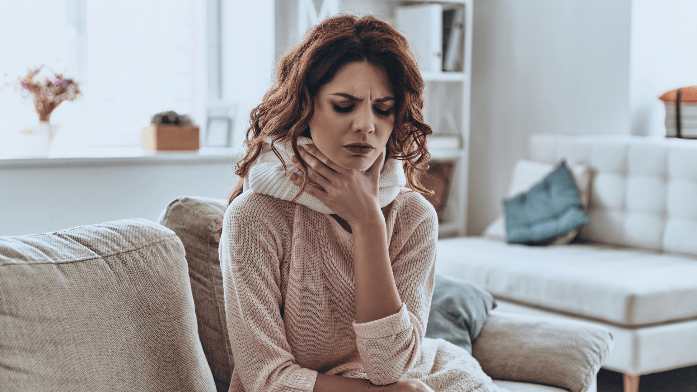 woman with red hair clutching her sore throat while sitting on couch