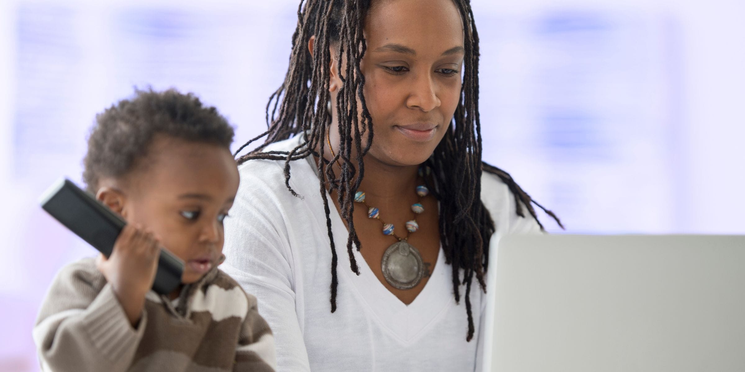 black mom working remotely on her laptop from home next to her son holding tv remote control