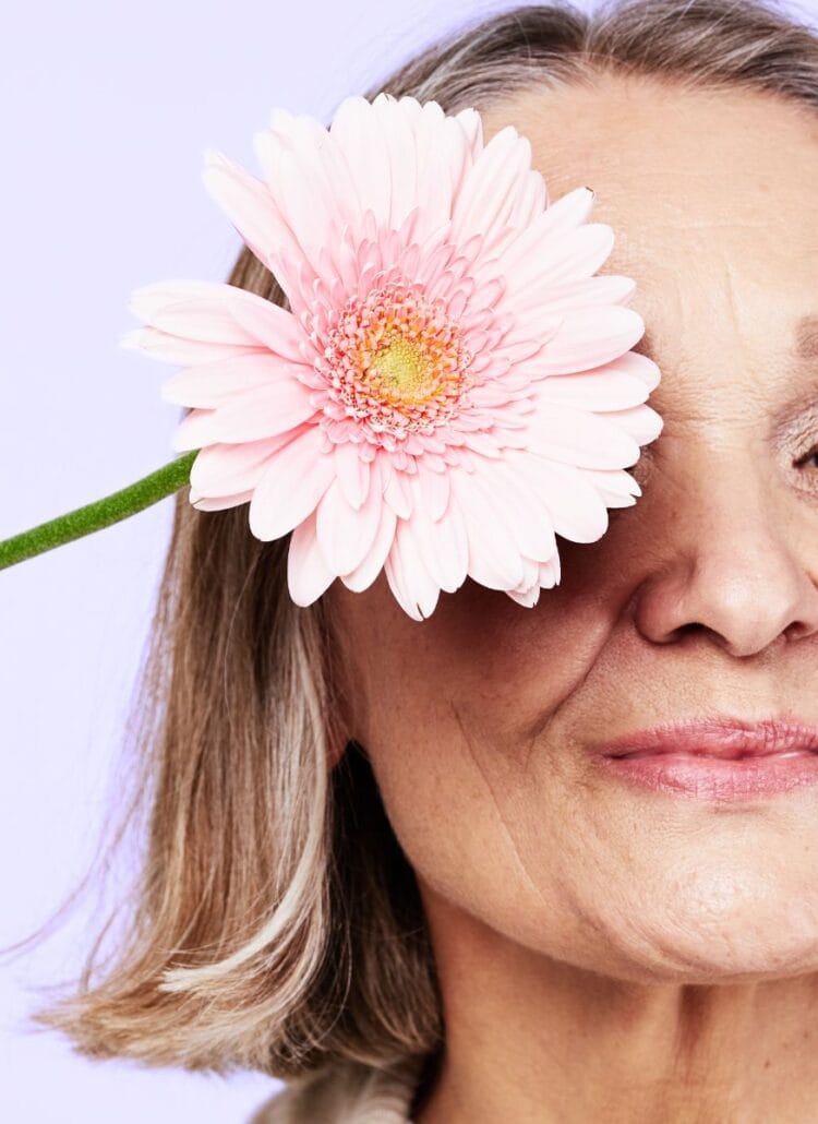 beautiful older woman holding pink daisy upt to her eye smiling