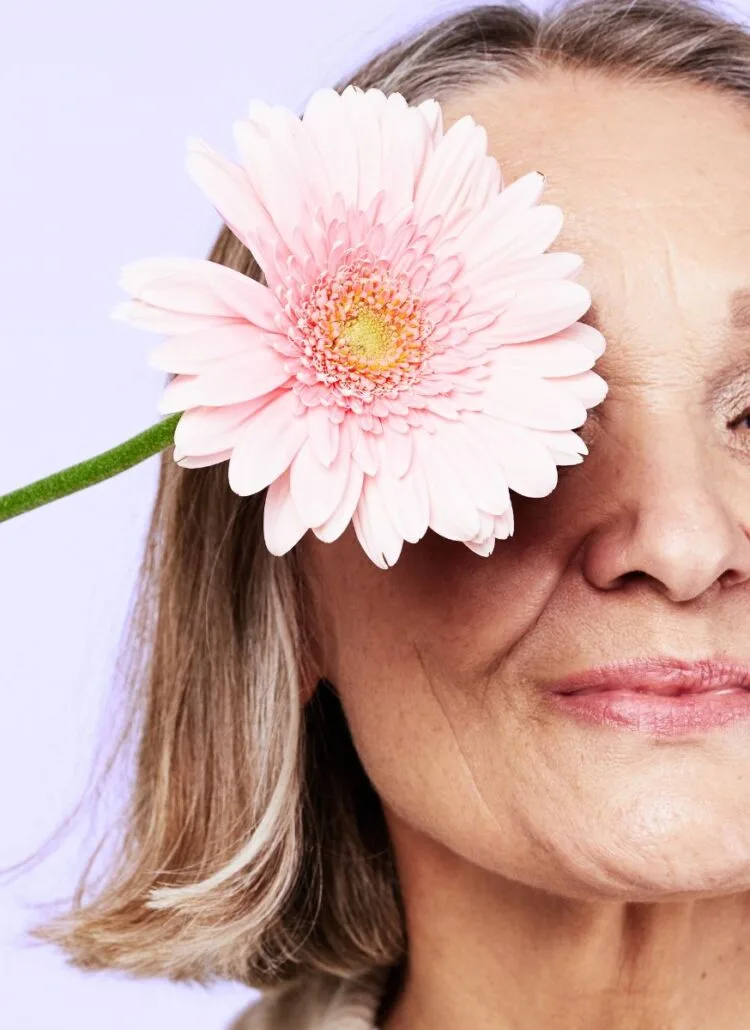 beautiful older woman holding pink daisy upt to her eye smiling