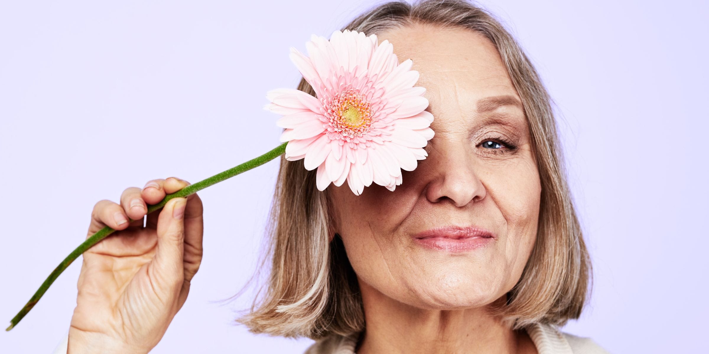 beautiful older woman holding pink daisy up to her eye smiling