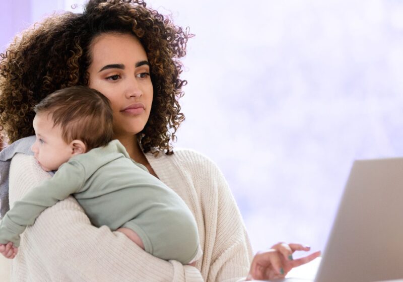 woman holding baby in green clothing while looking up parenting tips on laptop