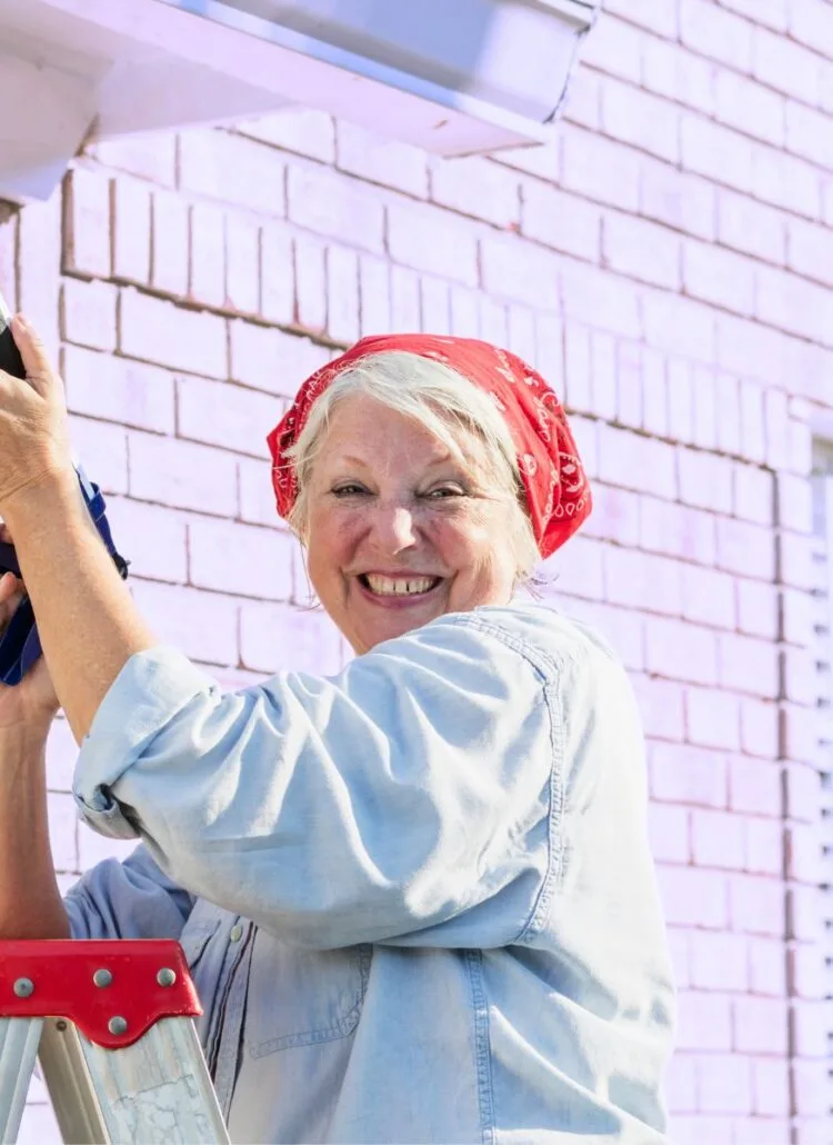 older woman doing home maintenance outside caulking exterior of house