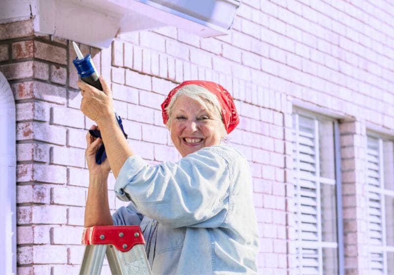 older woman doing home maintenance outside caulking exterior of house