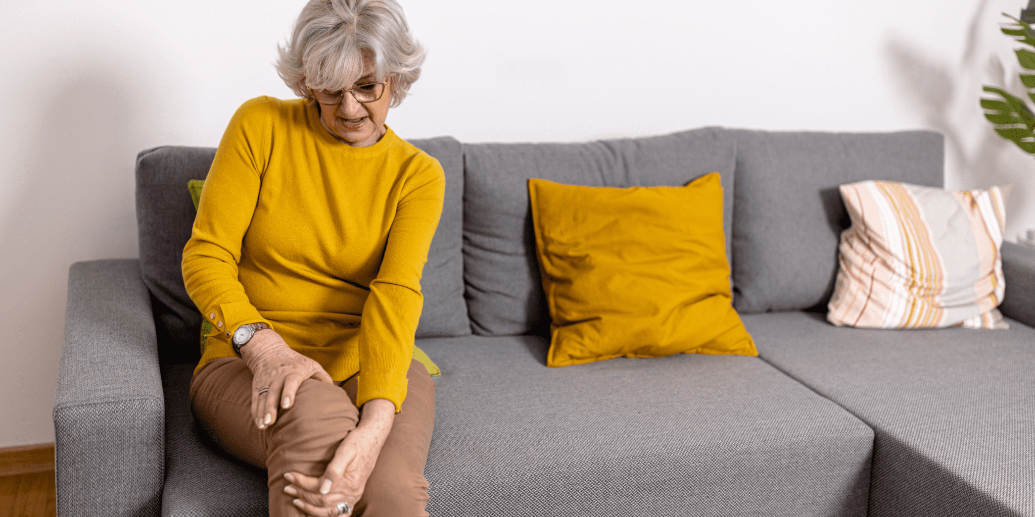 senior woman wearing yellow shirt sitting on gray couch holding knee hurting with chronic arthritis pain