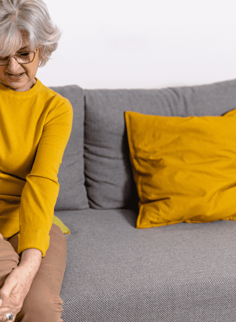 senior woman wearing yellow shirt sitting on gray couch holding knee hurting with chronic arthritis pain