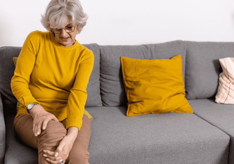 senior woman wearing yellow shirt sitting on gray couch holding knee hurting with chronic arthritis pain